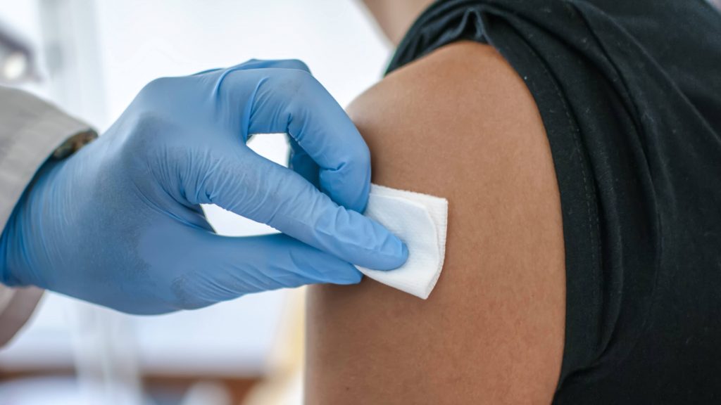 a white person's arm with a sleeve rolled up while a medical staff person, perhaps a doctor or nurse, wearing blue protective gloves wipes the arm with a cotton, preparing to administer a vaccine shot